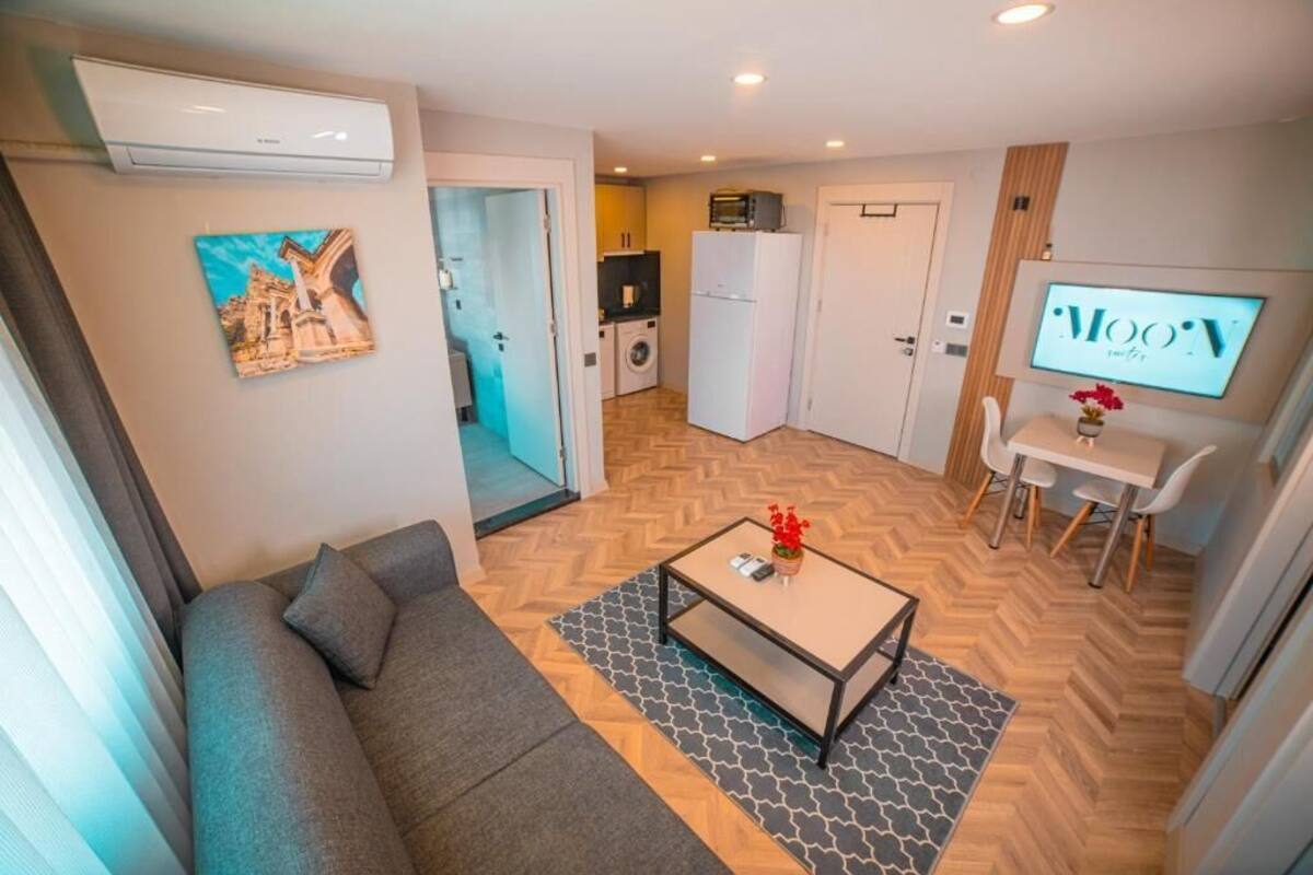 A modern living room with a sofa, coffee table, and a TV. There is a kitchen area with a microwave and refrigerator.