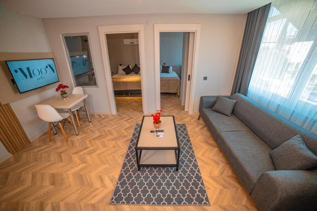 A modern living room with a sofa, a coffee table, and a TV mounted on the wall. There is a dining area with a table and chairs, and a hallway leading to a bedroom.