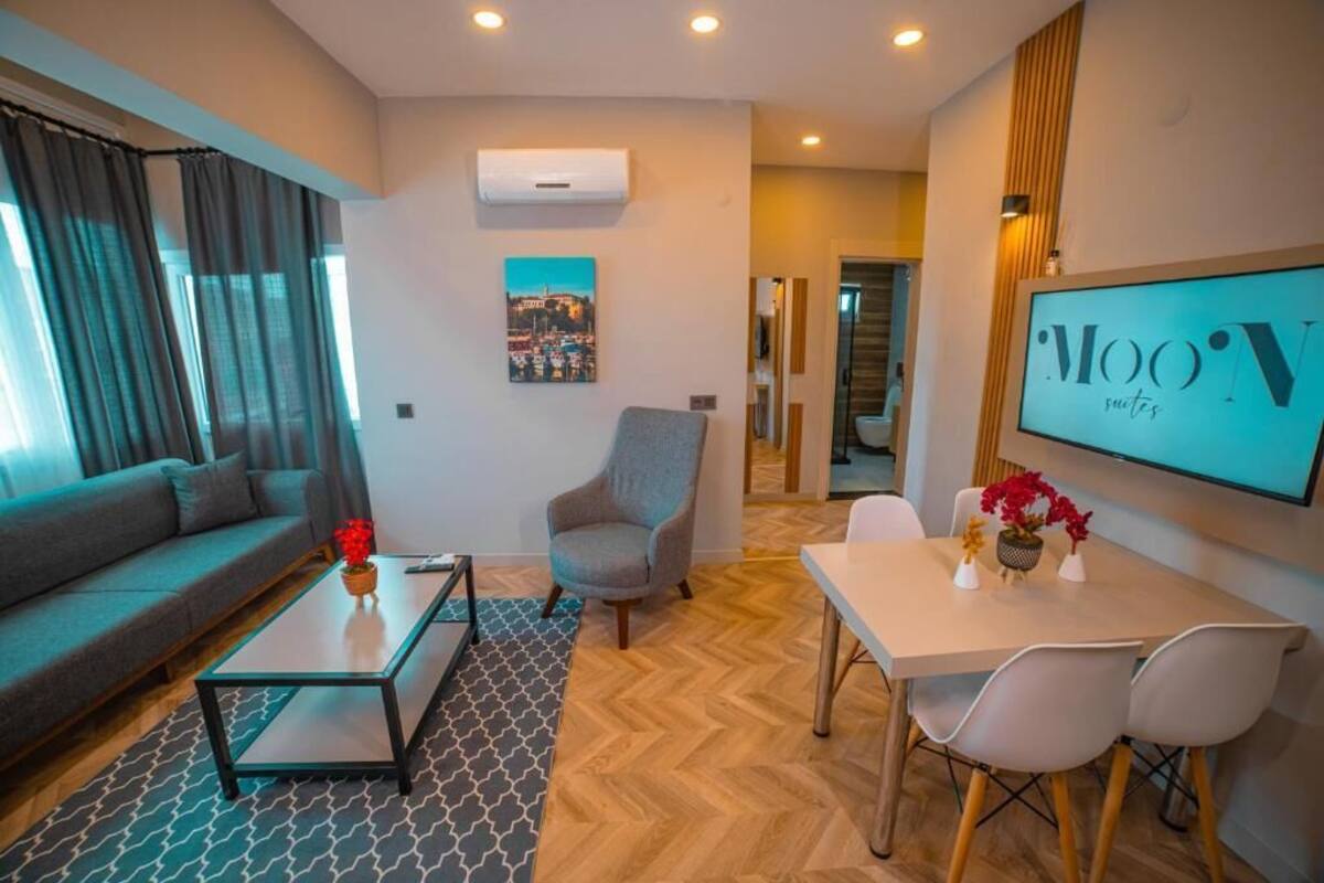 A modern living room with a sofa, armchair, coffee table, and dining area with chairs and a TV displaying 'MOON suites'.