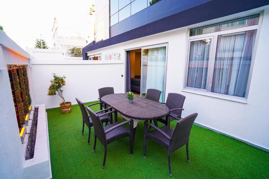 A patio with a round table and chairs, surrounded by artificial green turf, near a white building with a window and a door.