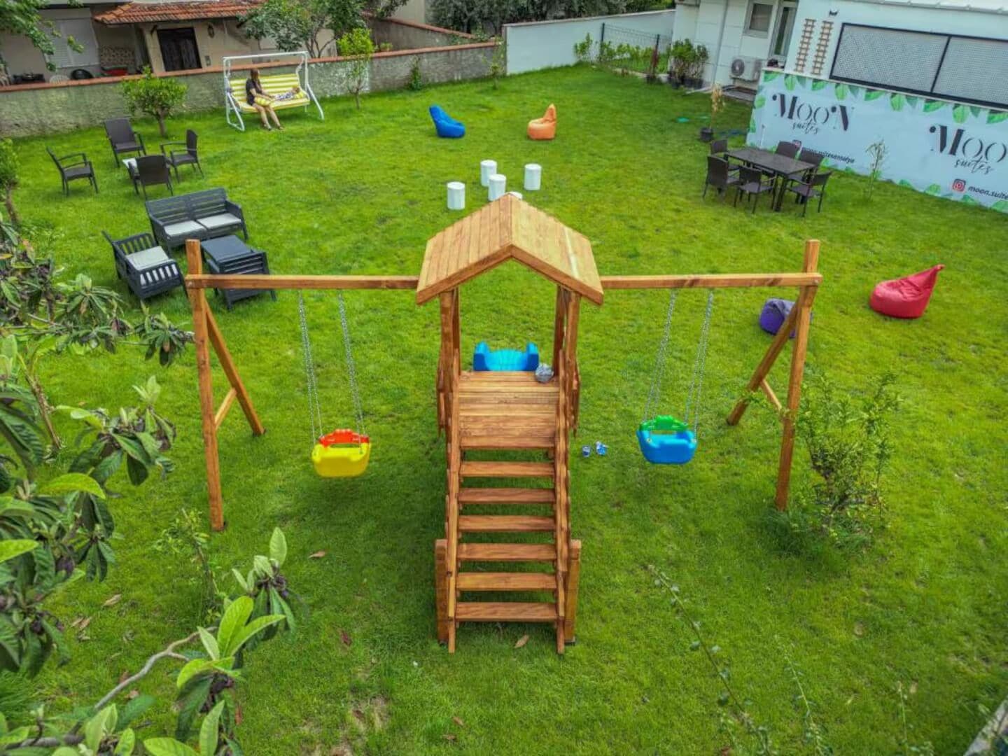 A wooden swing set with a ladder, surrounded by green grass, chairs, and bean bags.
