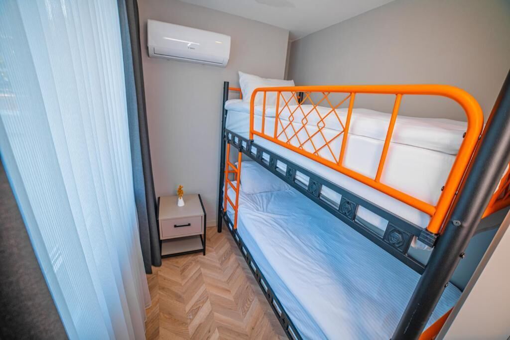 A bunk bed with an orange frame and white bedding, a small bedside table with a lamp, and an air conditioning unit on the wall.