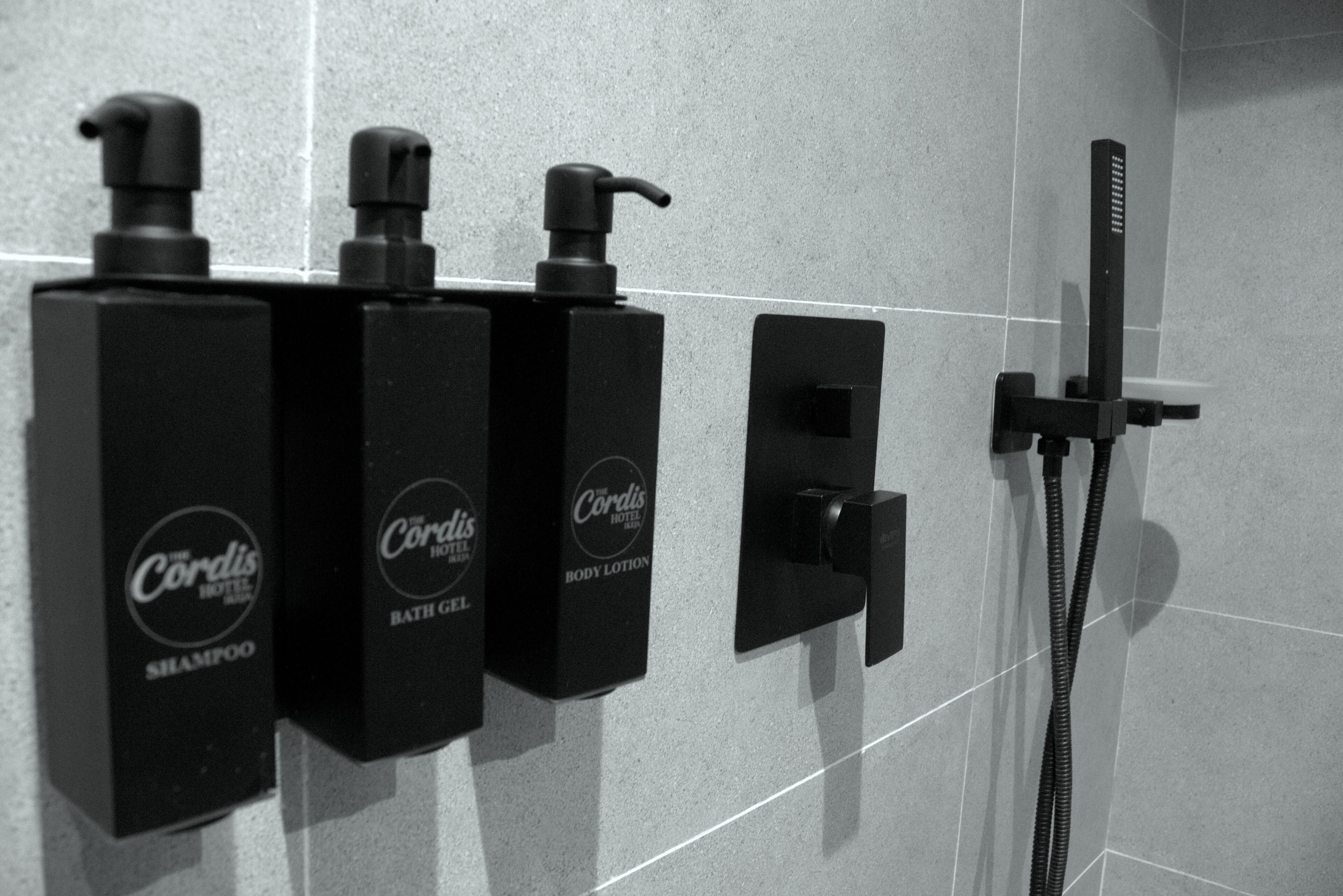 A bathroom with three black bottles of Cordis Hotel body lotion and shampoo, a black handheld showerhead, and a black faucet.