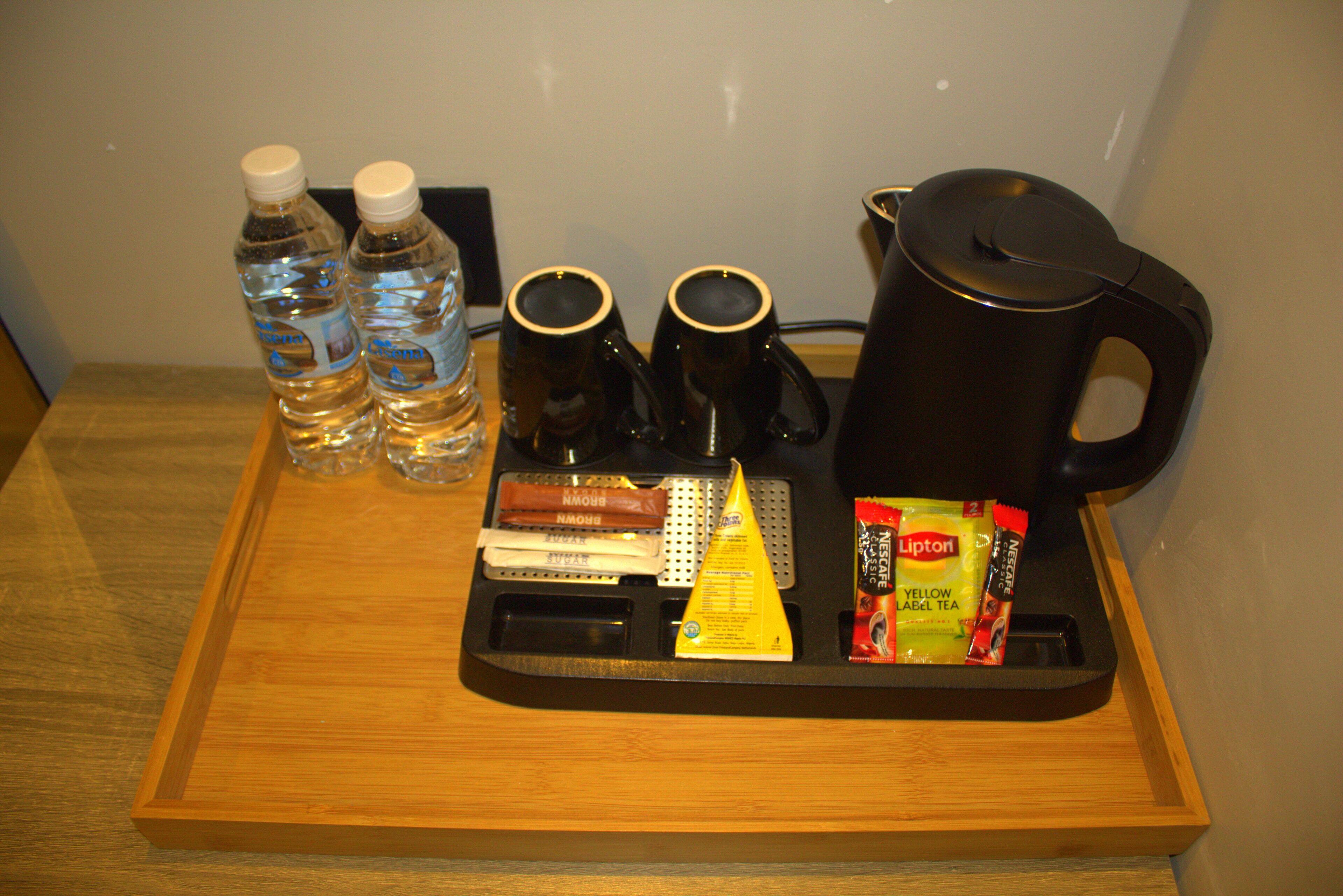 A tray with a coffee maker, two mugs, two bottles of water, a packet of tea, and a brown bag.