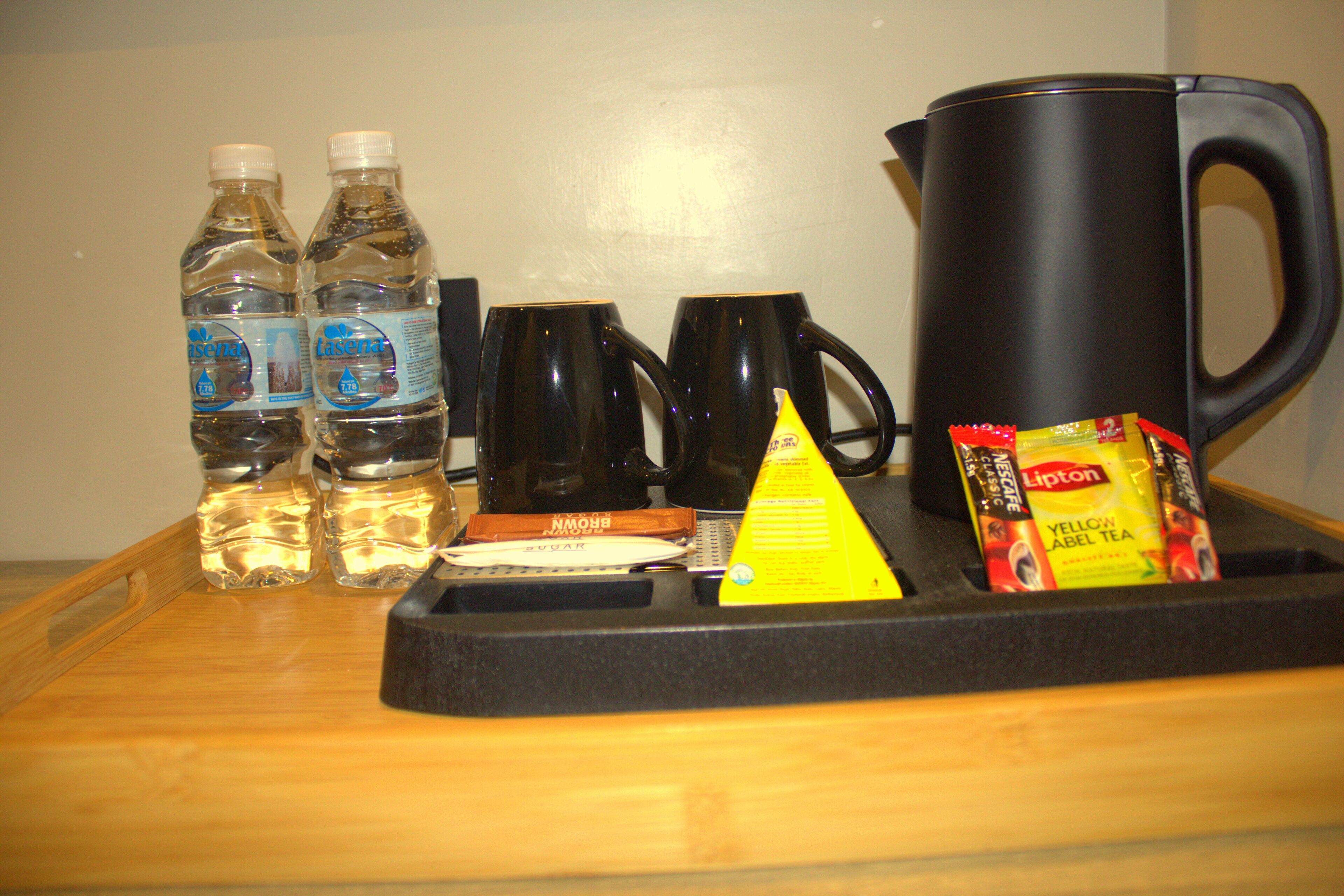 A tray with two bottles of water, two black mugs, a yellow triangular napkin, a black coffee pot, and Lipton tea packets.