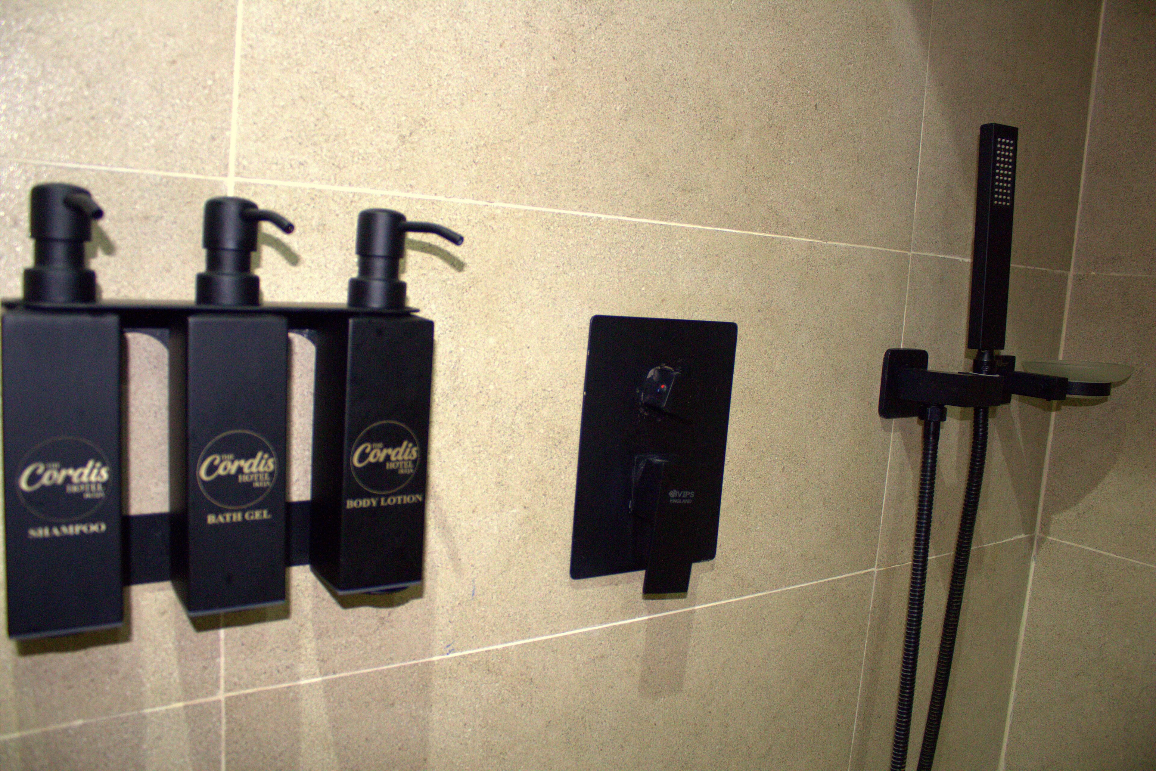 A bathroom with three bottles of Cordis body lotion, a handheld showerhead, and a wall-mounted faucet.