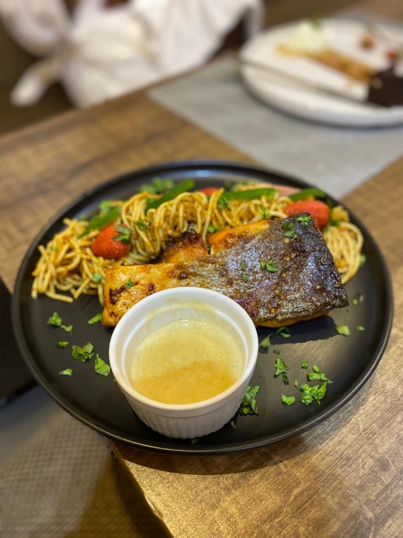 A plate with noodles, a piece of meat, and a small bowl of sauce on a table.