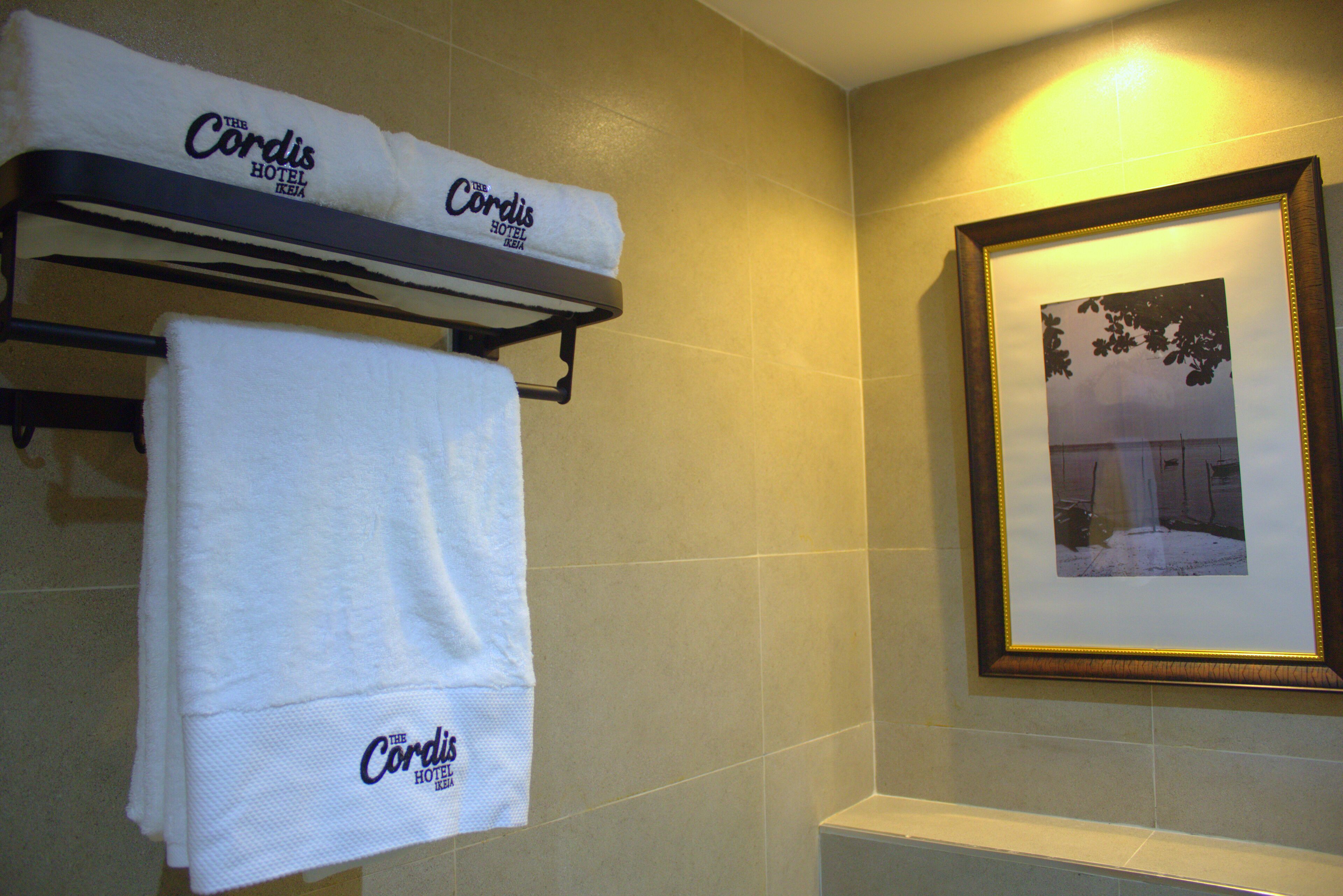 Une salle de bain équipée d’un porte-serviettes sur lequel sont posées deux serviettes, dont l’une porte le logo de l’hôtel Cordis, et d’un tableau encadré accroché au mur.