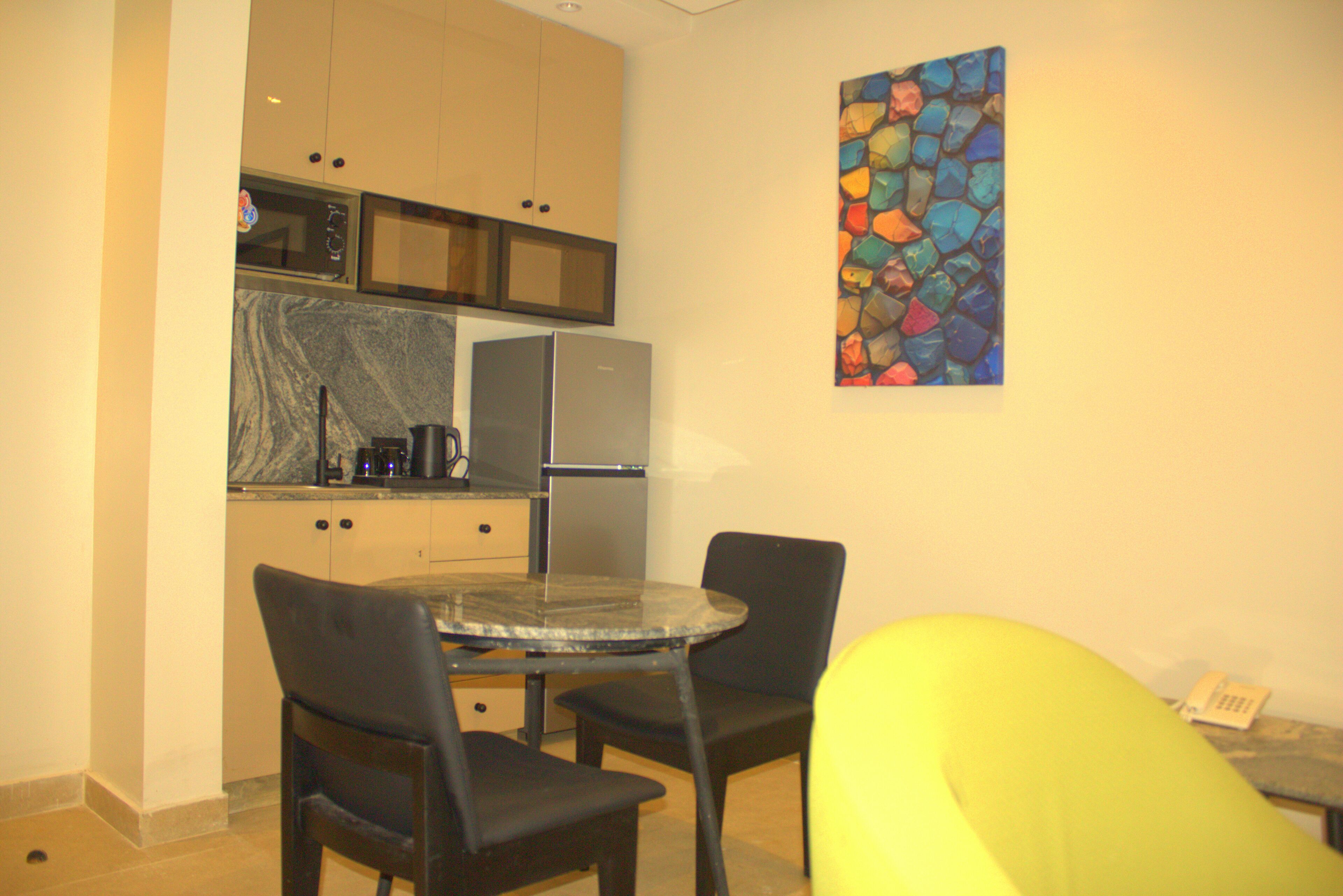 Una cocina compacta con microondas, estufa y refrigerador. Hay una pequeña mesa de comedor con dos sillas y un cuadro abstracto de colores en la pared.
