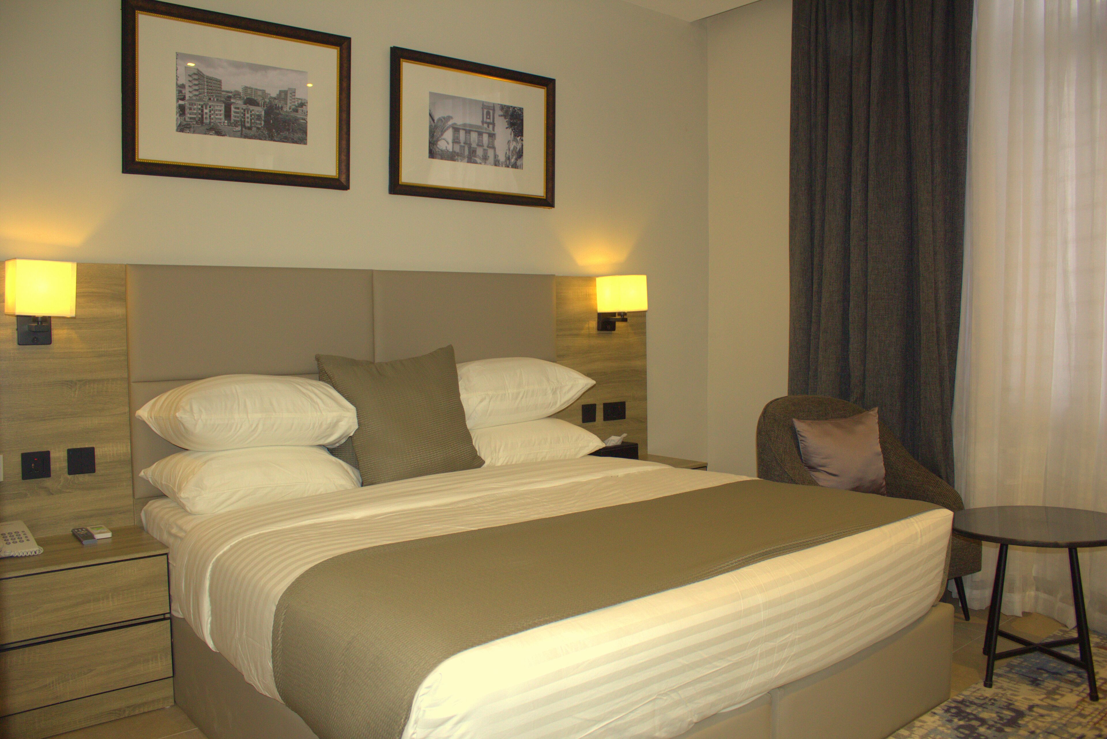 A hotel room with a large bed, two bedside lamps, a chair, a small table, and two framed pictures on the wall.