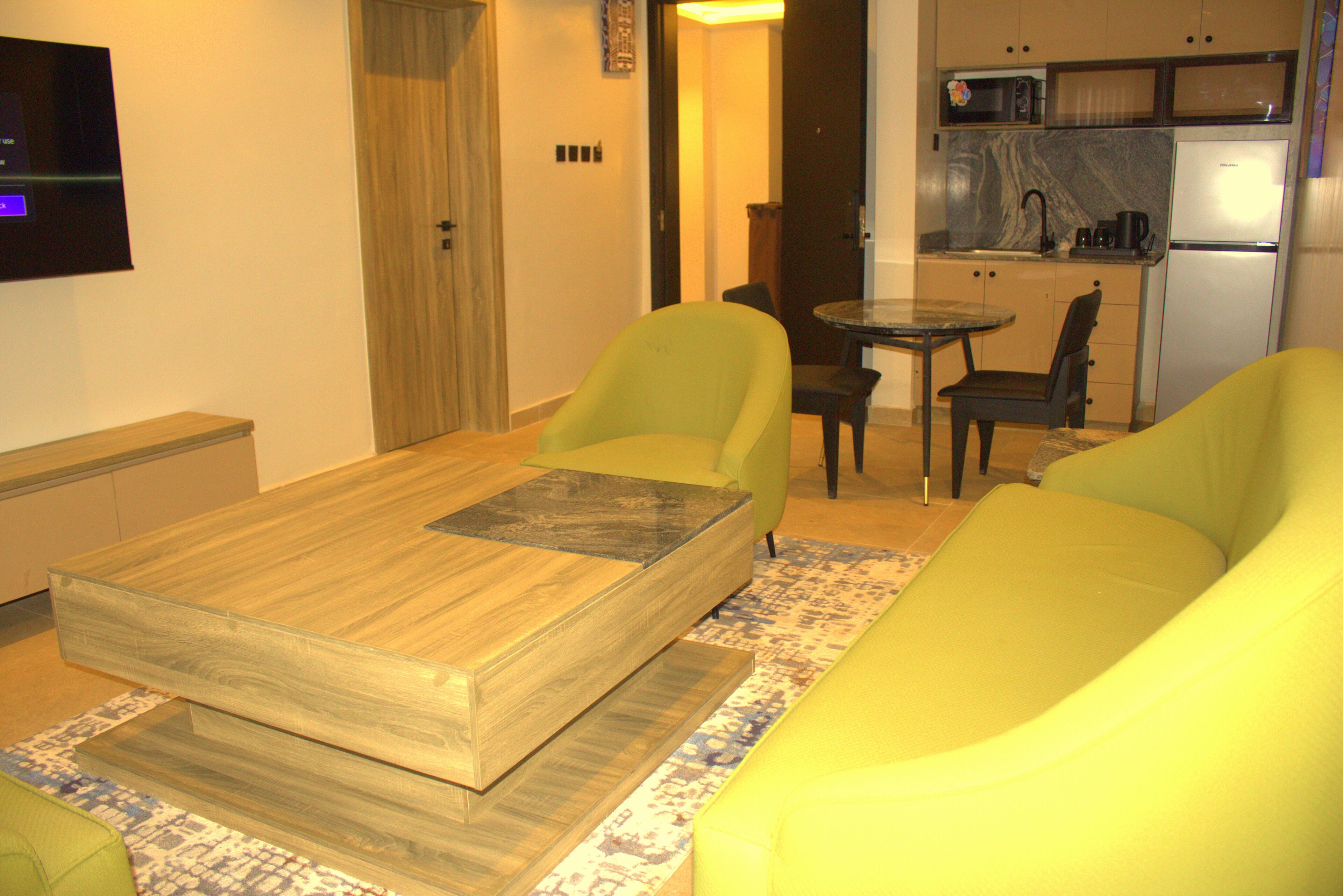 A modern living room with a wooden coffee table, two green armchairs, a small dining table, and a kitchen area with a refrigerator and microwave.