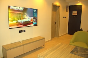 Una habitación de hotel moderna con un televisor grande montado en la pared, un banco de madera, una silla verde y una puerta con un letrero.