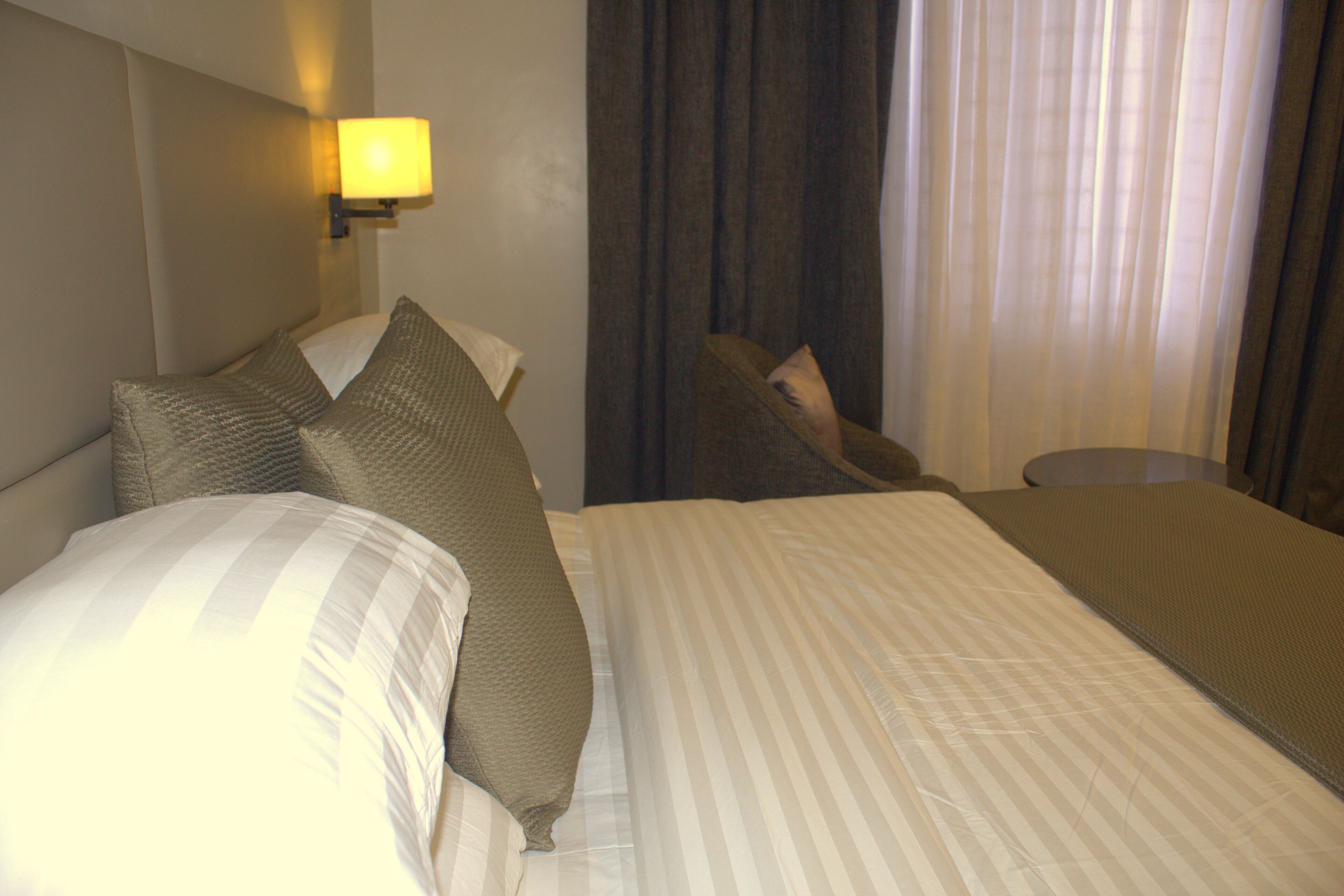 A hotel room with a bed, pillows, a nightstand, a lamp, and a window with curtains.