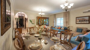 Un coin repas avec une grande table dressée pour six personnes, entourée de chaises en bois. Un lustre est suspendu au-dessus de la table, et un canapé et des fauteuils sont visibles à l’arrière-plan.