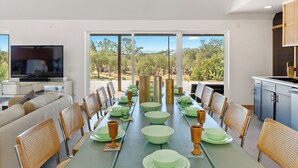 Een eetgedeelte met een lange tafel gedekt voor zes personen, omringd door rieten stoelen, met een televisie en keuken op de achtergrond.