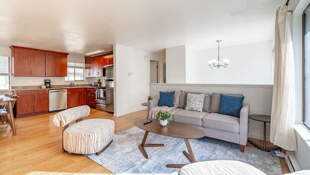 Una sala de estar moderna con sofá, reposapiés y mesa de centro, una cocina con armarios de madera y electrodomésticos de acero inoxidable, y un amplio ventanal con cortinas.