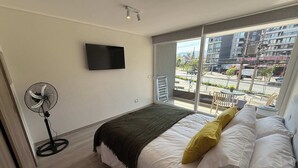 Une chambre à coucher comprenant un lit, une télévision, un ventilateur et un balcon avec vue sur des immeubles et une rue.
