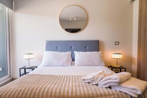 Studio bedroom with a blue-gray tufted headboard, soft layered bedding, and matching bedside lamps beside a bright window. - Modern Studio in Las Condes with Gym & BBQ (Las Condes)