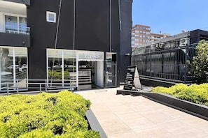 Modern apartment entrance with glass doors, landscaped planters, and a sunlit tiled walkway. - Modern Studio in Las Condes with Gym & BBQ (Las Condes)