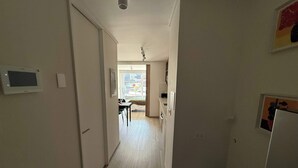 Un couloir étroit, avec une porte sur la gauche, une fenêtre au fond, et une petite table entourée de chaises, visible au loin.
