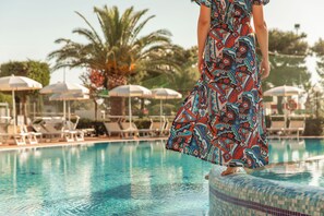 A person in a patterned dress standing at the edge of a pool with lounge chairs and umbrellas in the background. - Residence camping Atlantide (Capitolo)