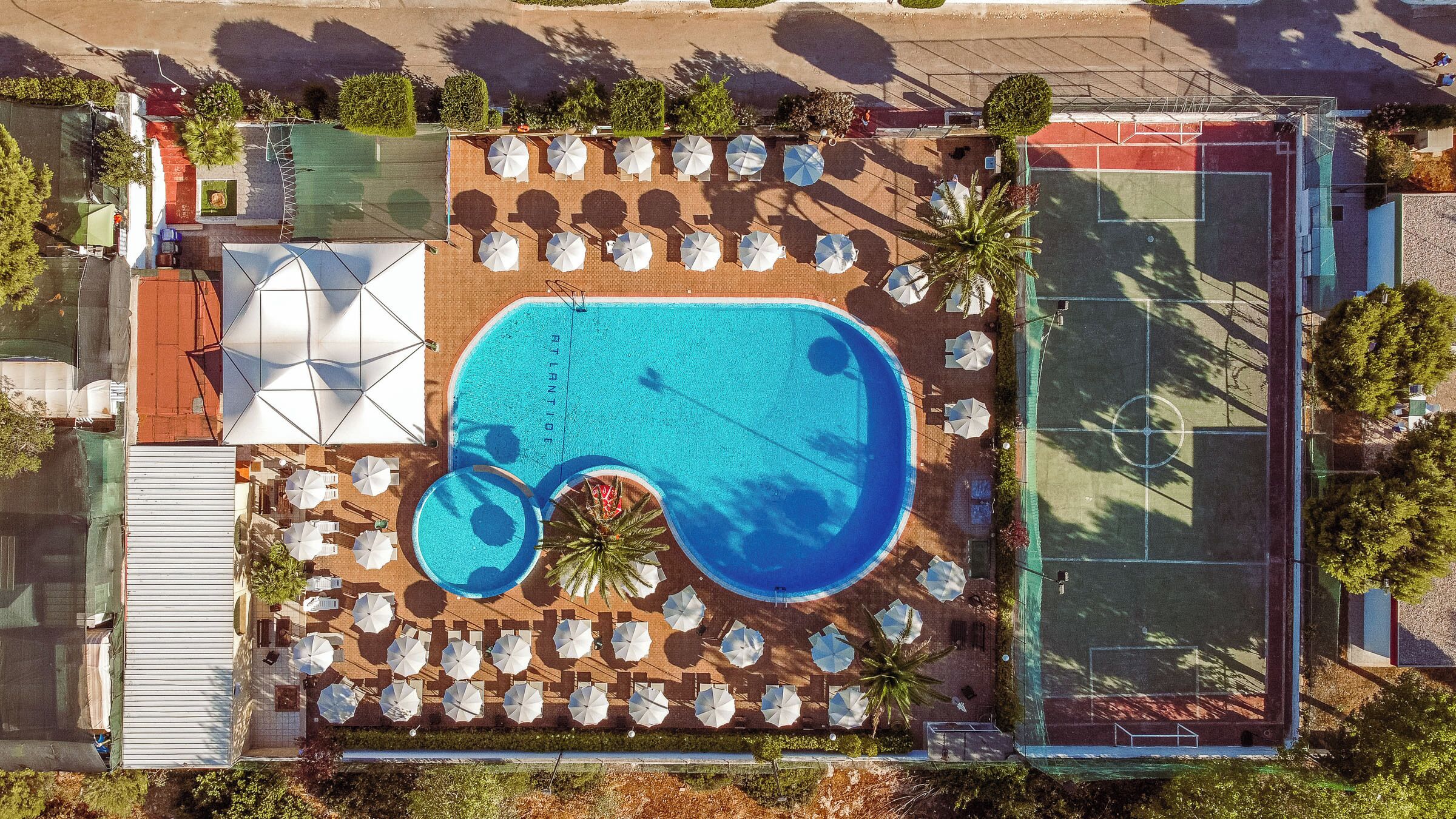 An aerial view of a pool area with multiple sun loungers and umbrellas, a tennis court, and a small garden area.