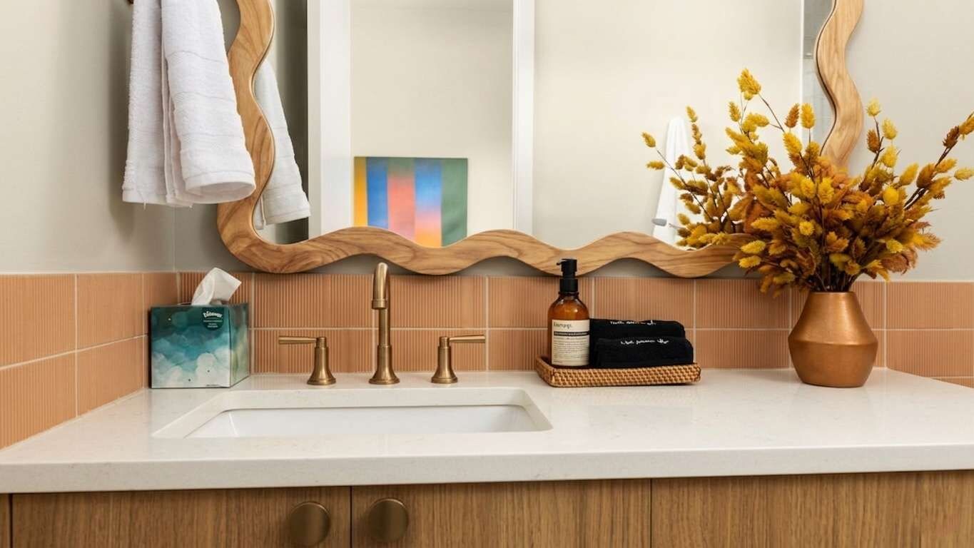 A bathroom with a sink, a mirror, a towel rack, a bottle, and a vase with dried flowers.
