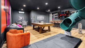 Un espacio lounge moderno con mesa de billar, asientos cómodos, un bar y un tobogán verde grande.