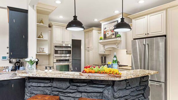 Una cocina con una isla grande, dos lámparas colgantes, un refrigerador y una pared de acento de piedra.