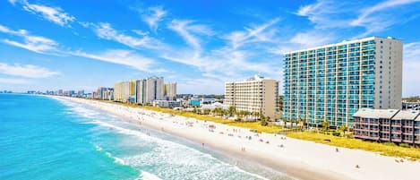 Un hotel frente a la playa con varios edificios altos, una playa de arena y un cielo azul despejado.