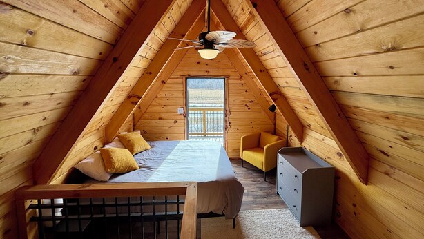 Une chambre mansardée chaleureuse, avec un plafond en pente, un lit agrémenté d’oreillers jaunes, une petite chaise, une commode et une fenêtre donnant sur l’extérieur.