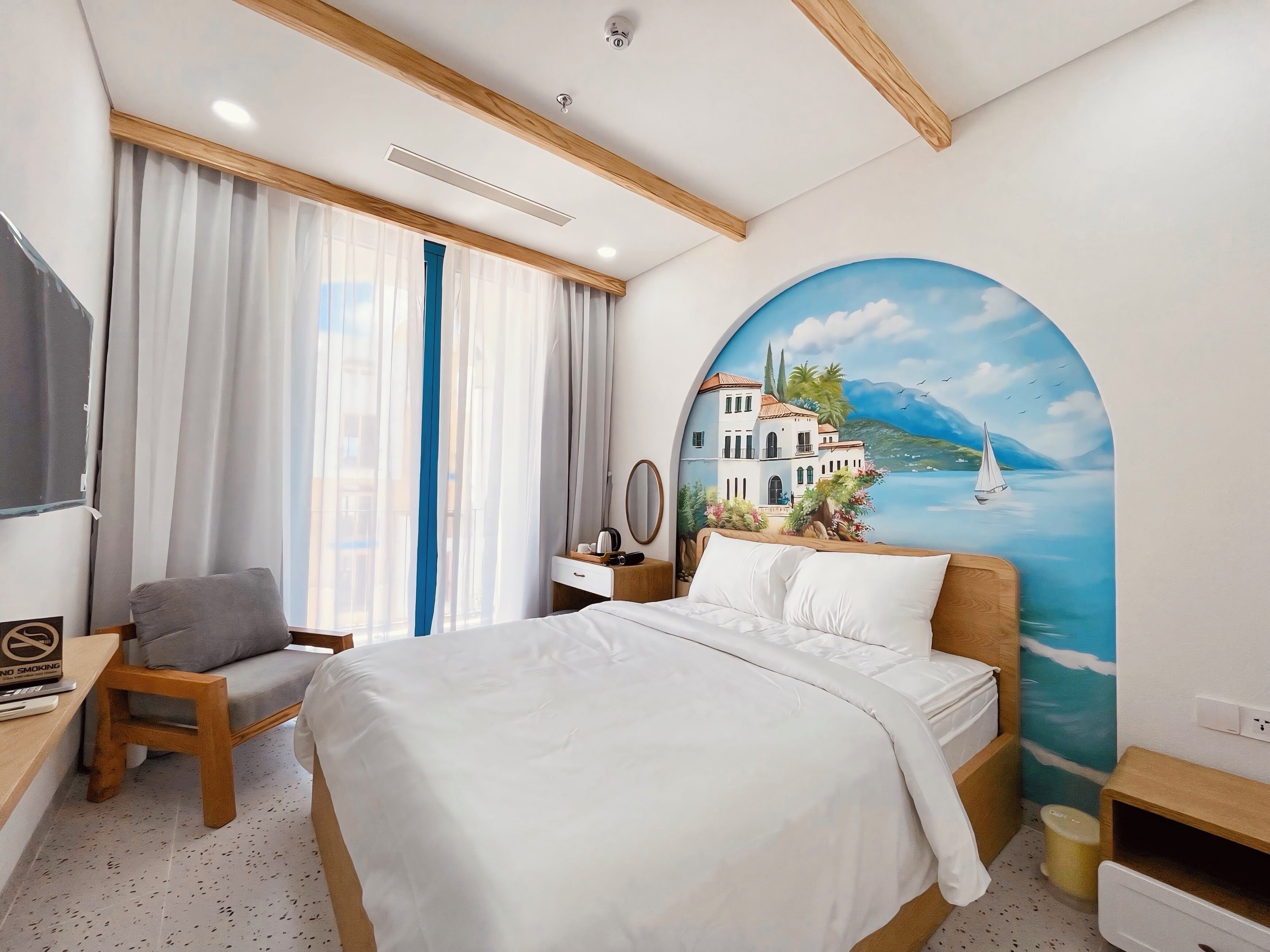 Habitación de hotel con cama, escritorio, silla, mesita de noche y un cuadro con una escena costera.