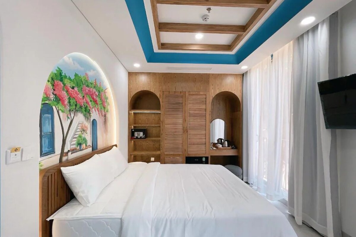 Una habitación de hotel con una cama, televisor, un cuadro, estanterías de madera y una ventana con cortinas.