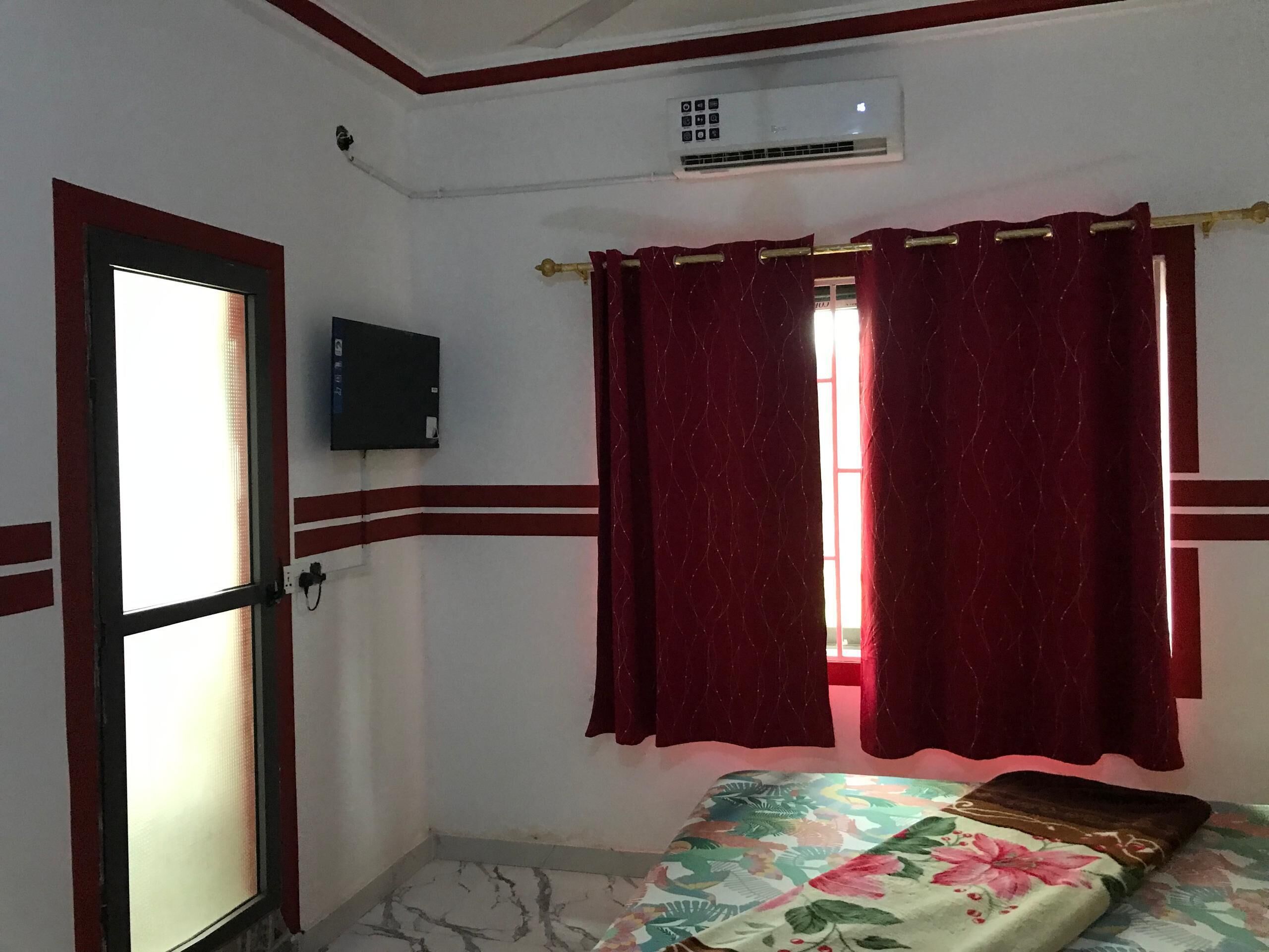Un dormitorio con cama, ventana con cortinas rojas, unidad de aire acondicionado y televisor instalado en la pared.