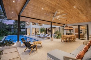 A modern outdoor living area with a pool, lounge chairs, a dining table, and a ceiling fan.