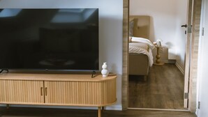 Een moderne woonkamer met een flatscreen televisie, een houten kast en een slaapkamer die door een open deur zichtbaar is.