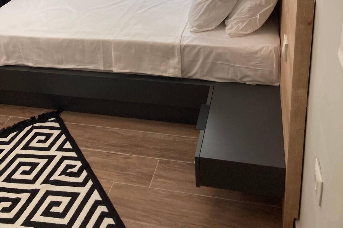 A neatly made bed with white linens and pillows, a wooden headboard, and a geometric patterned rug on a wooden floor.