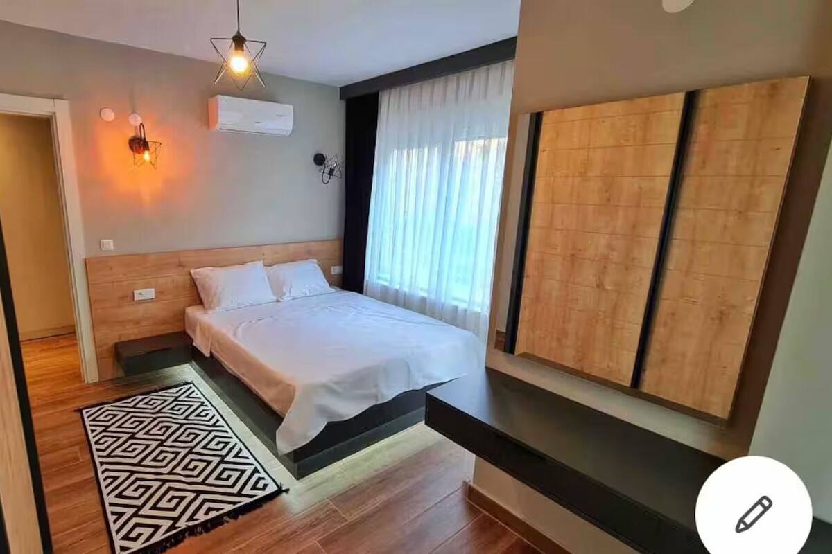 A modern bedroom with a wooden headboard, a bed with white bedding, a geometric-patterned rug, and a wall-mounted air conditioner.