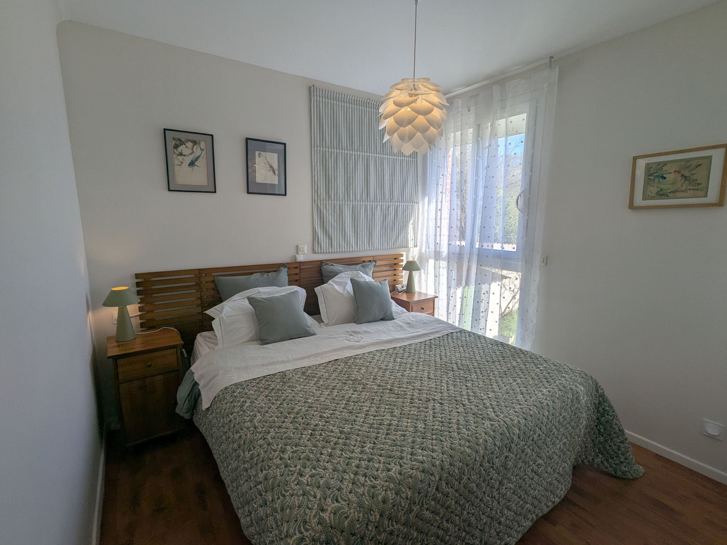 Une chambre à coucher avec un grand lit, une tête de lit en bois, deux tables de chevet avec des lampes, un luminaire suspendu et une fenêtre avec des rideaux légers.