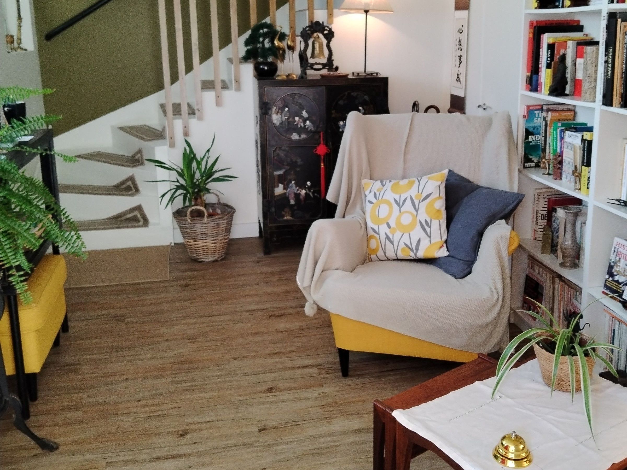 Un salon avec un fauteuil jaune, un plancher en bois, une bibliothèque, un escalier et une plante en pot.