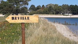 Un letrero de madera con 'REVILLE' y 'Jonville' indica el camino hacia una playa con aguas tranquilas y árboles al fondo.