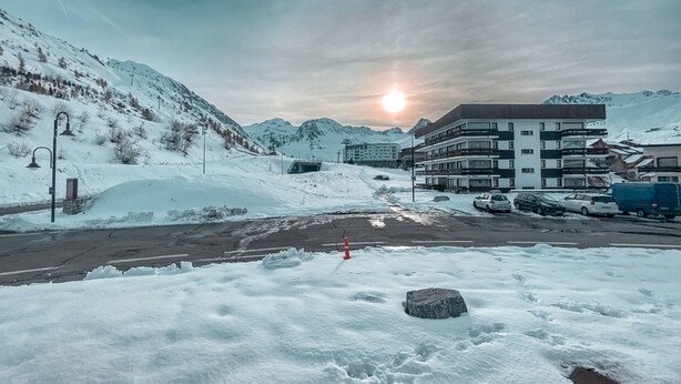 Paisaje nevado con un edificio de varios pisos y balcones, un área de estacionamiento con varios coches y una montaña al fondo.