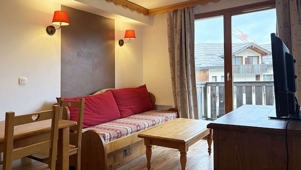 Une pièce chaleureuse avec un banc en bois, un canapé avec des coussins rouges, une petite table et un téléviseur posé sur un meuble en bois.