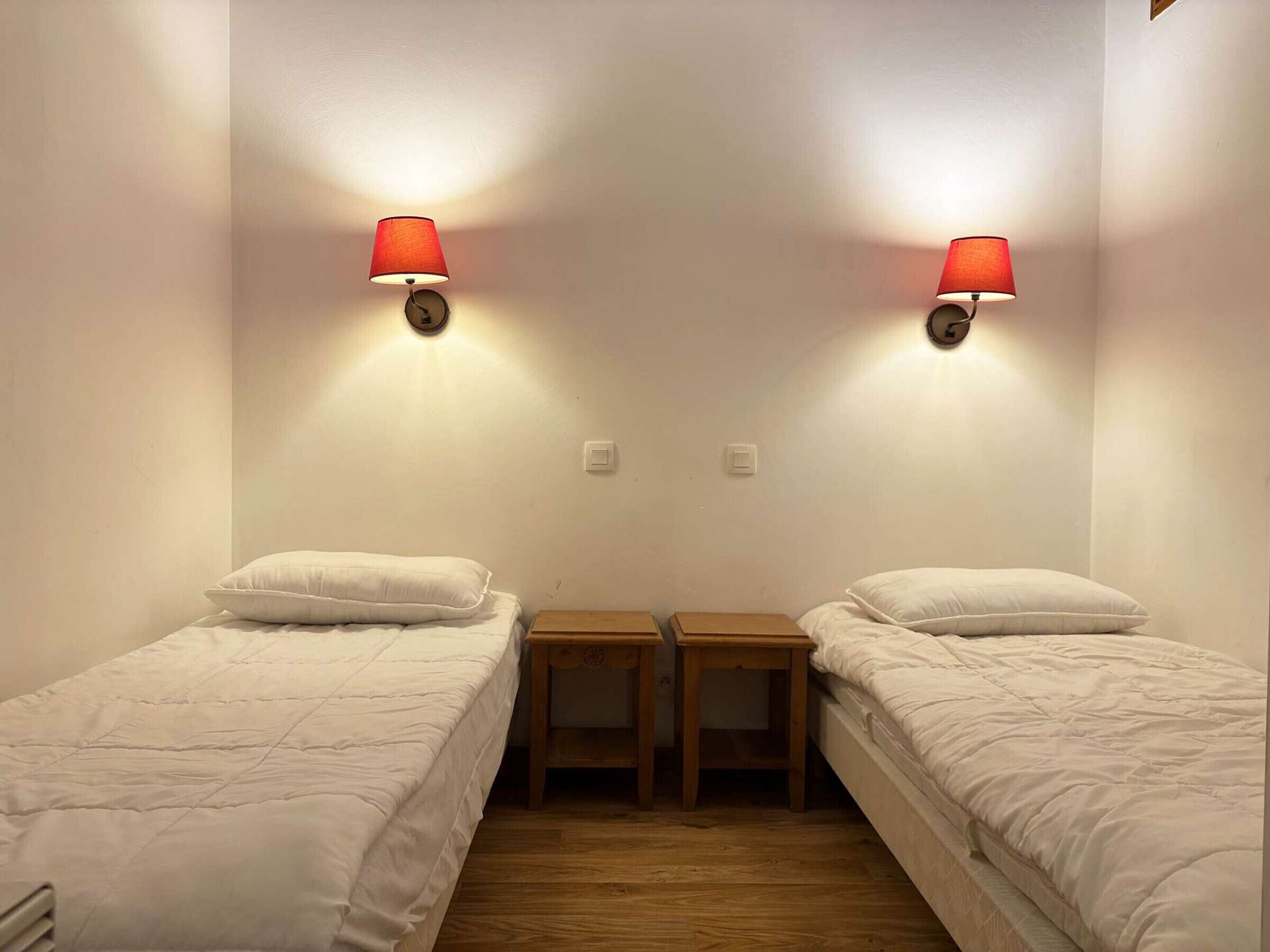 A room with two single beds, each with a wooden nightstand, and two wall-mounted lamps.