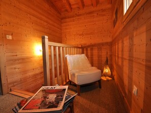 A cozy wooden cabin room with a white chair, a magazine on the floor, and a wall-mounted light fixture.