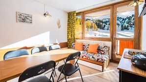 A room with a wooden table, chairs, a sofa, and a bed. There is a window with a view of trees and mountains. - Renovated Studio Cabin - On the Slopes at Plagne Centre (La Plagne-Tarentaise)