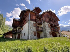A multi-story wooden building with balconies, stone pillars, and a mountainous backdrop. - Bright apartment near slopes, with parking (Serre Chevalier Chantemerle)