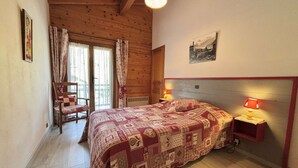 A bedroom with a wooden ceiling, a bed with a patterned quilt, a chair, a bedside table with a lamp, and a window with curtains. - 3-room apartment in Praz-Sur-Arly, 6 people, parking, near slopes (Praz-sur-Arly)