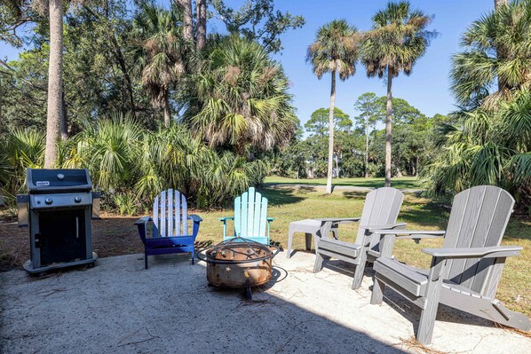 A patio with Adirondack chairs, a grill, and a fire pit surrounded by palm trees.