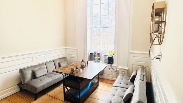 A spacious room with a wooden floor, a large window, a chandelier, and a sofa. - Liz Room in downtown Exeter next to PEA (Exeter)